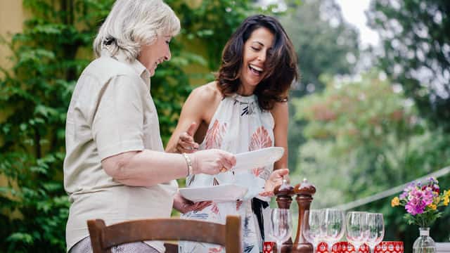 Older Mother and Daughter laugh and set outdoor dining table with wine glasses