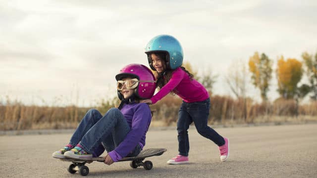 a girl sitting down on a skateboard while another girl is helping her push from the back