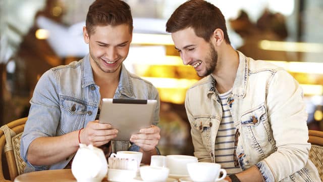 two men sitting down in a cafe smiling brightly while looking at a tablet