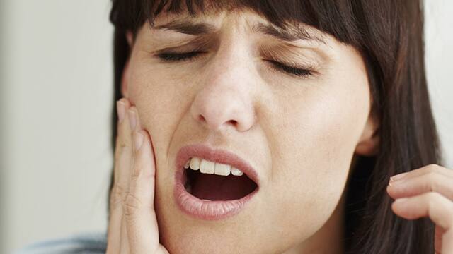 A brown hair woman with gum pain, using one hand to place pressure on the affected cheek.