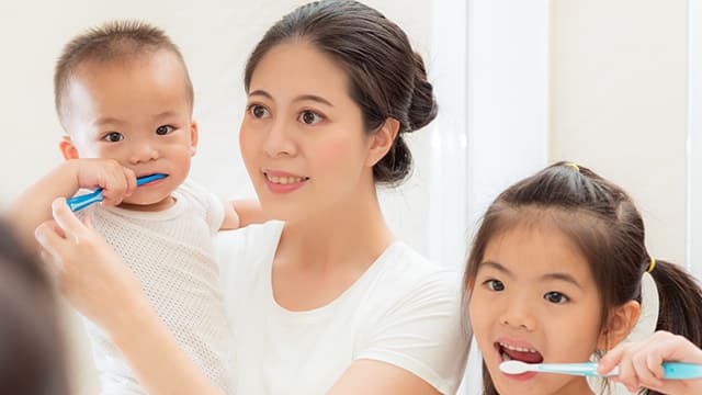 A mom helping her boy toddler to brush his teeth, while her young daughter brushes her own teeth by their side.