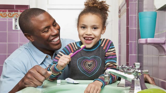 a father smiling while looking at his daughter brushing her teeth with Colgate toothpaste
