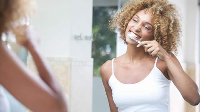 Woman with curly hair happily brushing her teeth in a bathroom mirror