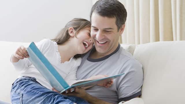 a father reading a book to the daughter sitting on her father lap while smiling