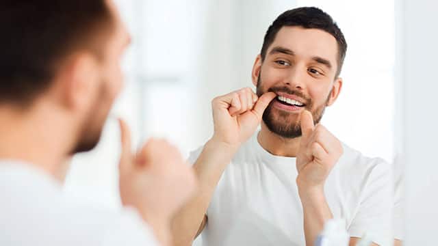 A man with dental floss cleaning teeth at a mirror in a bathroom