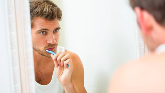 Young man in white tank top brushing his teeth in front of a bathroom mirror