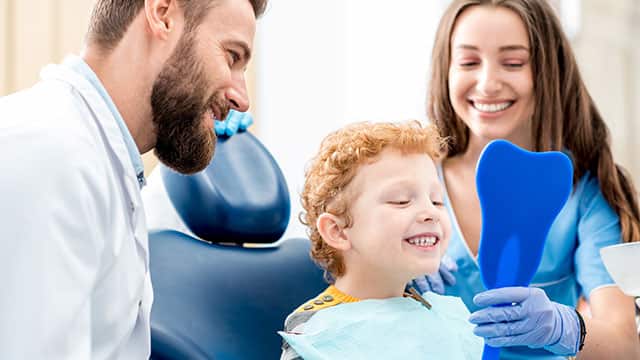 A young boy looking at the mirror with toothy smile sitting on the chair with dentist and assistant at the dental office