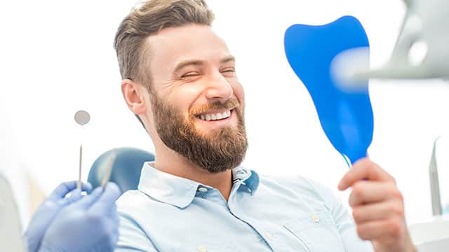 male patient looking at his smile with a hand mirror while sitting at the dental office