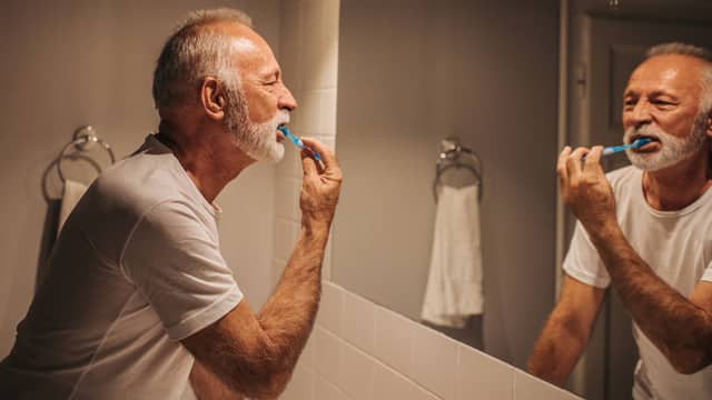 An older man with a beard brushing his teeth while looking in the bathroom mirror