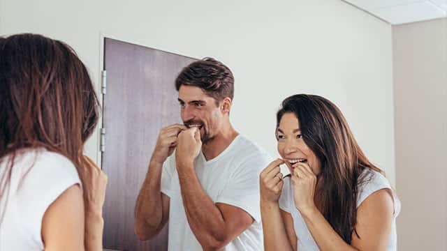 Couple using dental floss to clean their teeth in front of the mirror