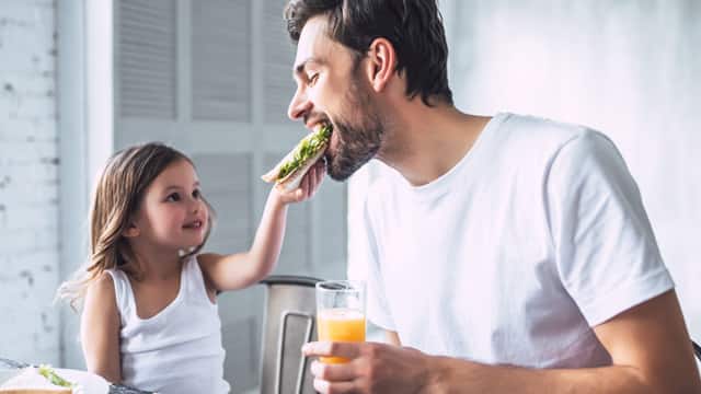 Young girl in a white shirt feeding a sandwich to her father in a white shirt