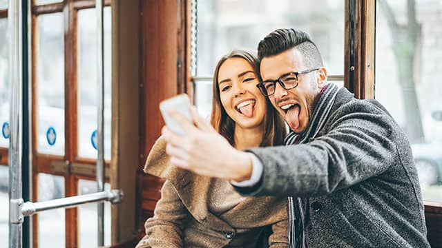 Happy young couple taking a selfie while sticking their tongue out using mobile smartphone camera inside the bus