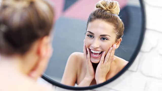 A young woman is looking into a bathroom mirror while touching her jaw and smiling