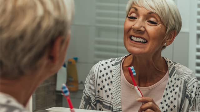 A senior woman examines her gums while brushing her teeth in a mirror