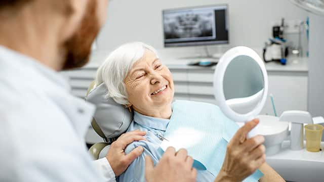 An elderly woman looking at her teeth as the dentist stands besides her