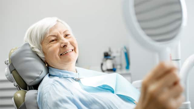 Senior woman in a dental chair smiling while looking at her dentures into a handheld mirror