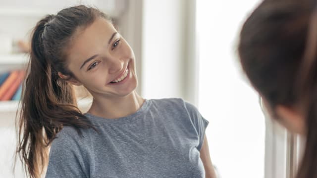 Young woman in a gray shirt smiles while looking at herself in a mirror