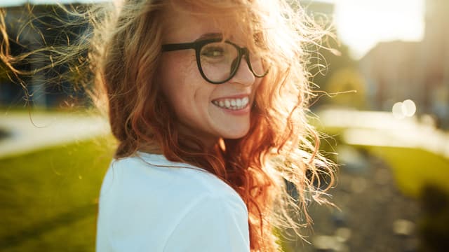 Young woman with red hair in thick, black-rimmed glasses smiling over her shoulder outdoors