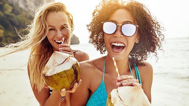Two laughing friends drinking from coconuts on a sandy beach