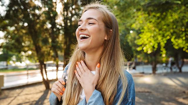 Cheerful young girl is laughing while putting her hand on her neck outside in a park