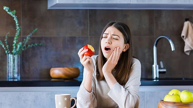 young woman is feeling toothache while eating apple in the kitchen