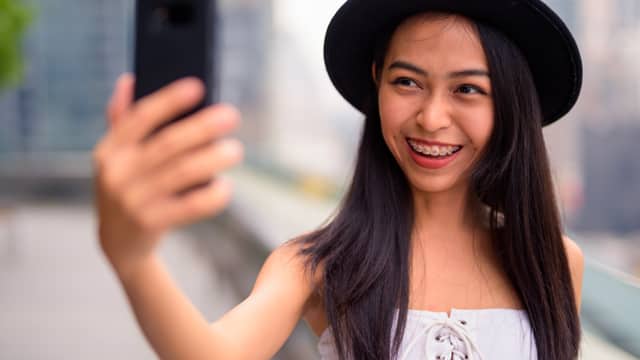 A young girl in a hat with braces taking a selfie outdoors