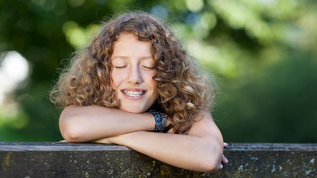 Smiling child with closed eyes and wearing braces is outdoors