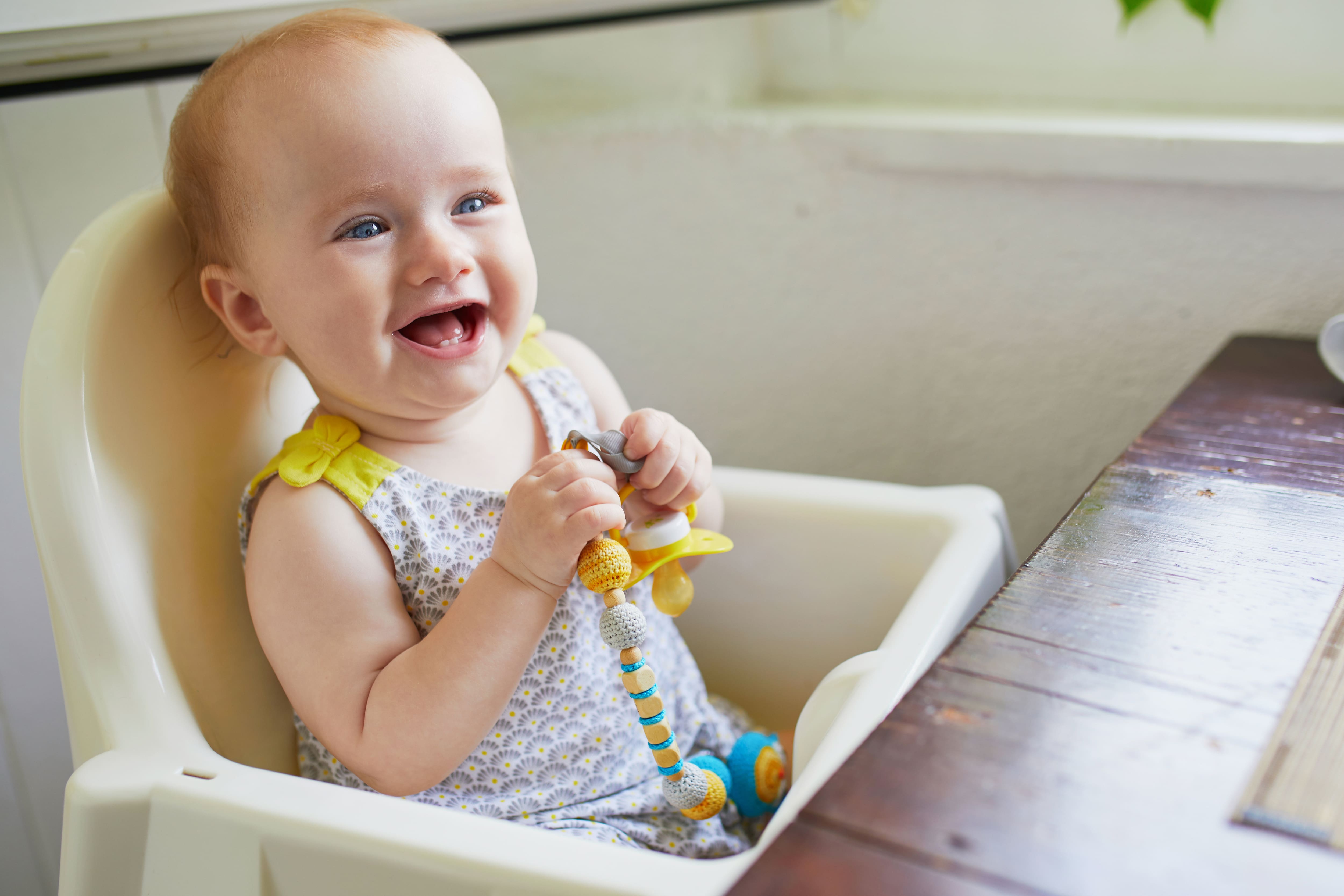 A little baby girl sitting in high chair and holding a pacifier while smiling