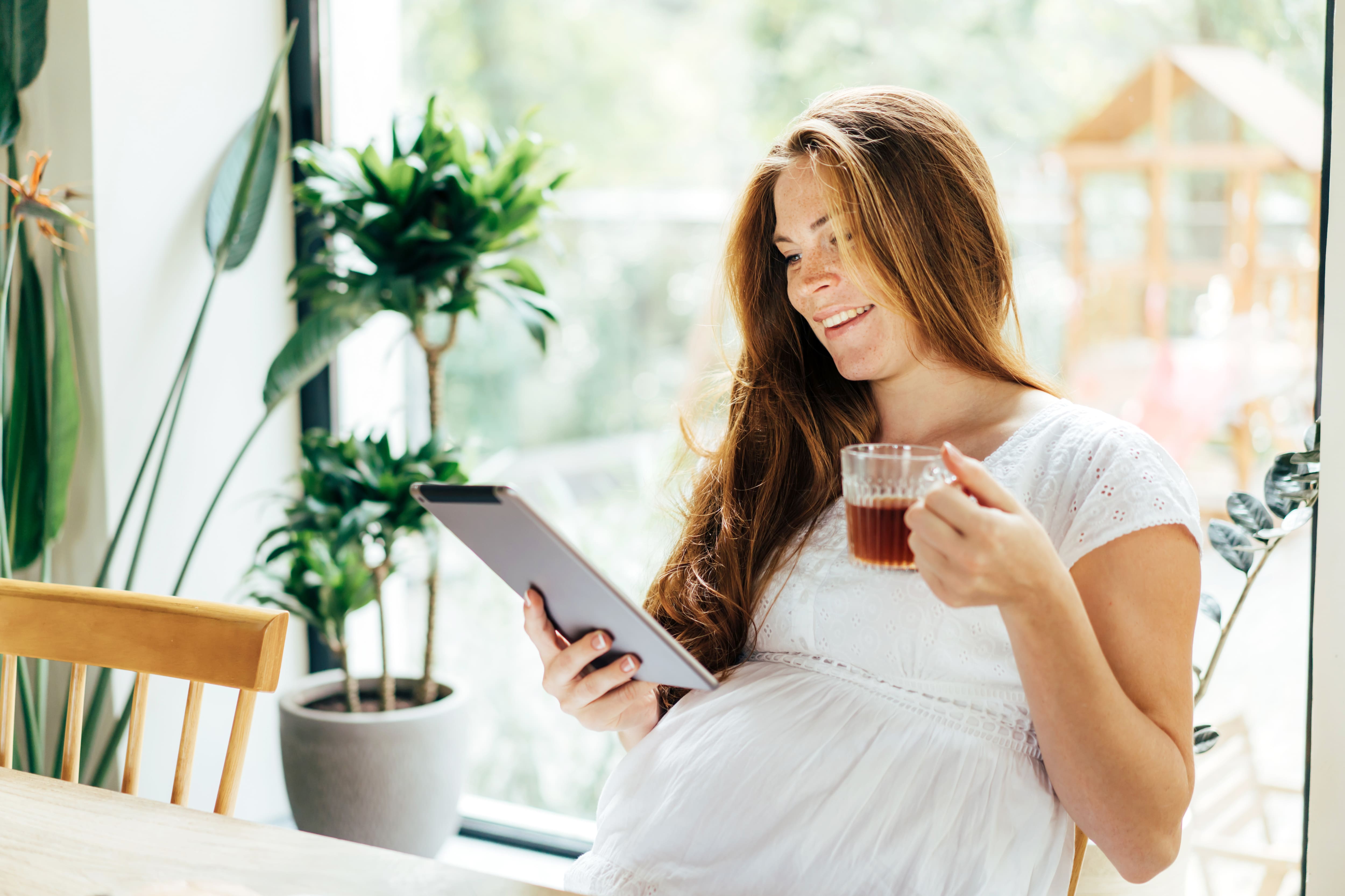 Pregnant woman drinks tea and looks at the screen of a computer tablet.