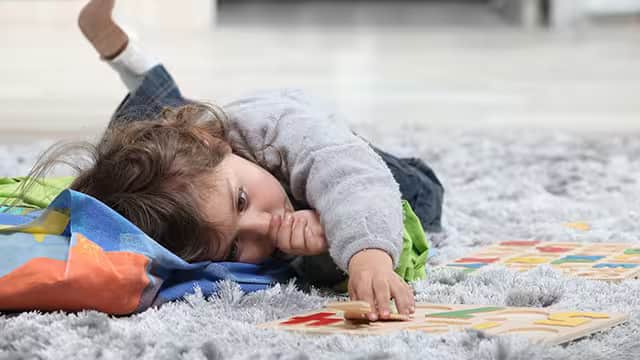 A toddler playing and sucking thumb A toddler playing and laying down on a rug while sucking their thumb