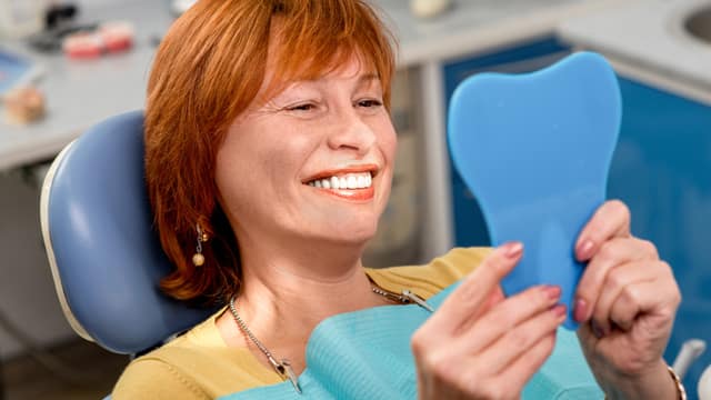 Older woman with red hair and smiling in a dental chair looking at her dentures in a handheld mirror