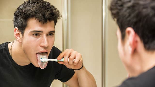 A young man is brushing teeth and tongue in front of the bathroom