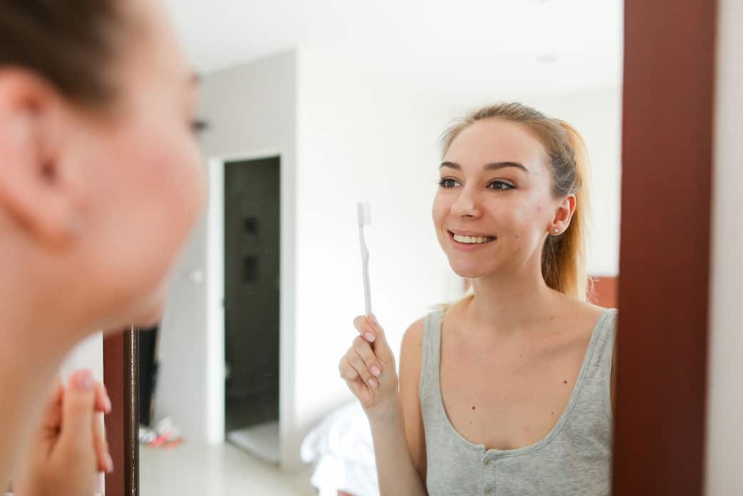 Young woman standing near mirror with tootbrush, wearing grey shirt.