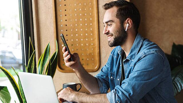 Photo of laughing caucasian man using earpod and cellphone with laptop while working in cafe indoors