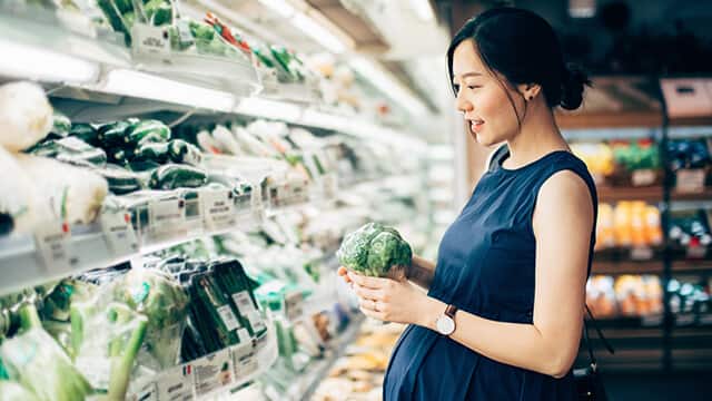 pregnant woman standing in the vegetable aisle while grocery shopping