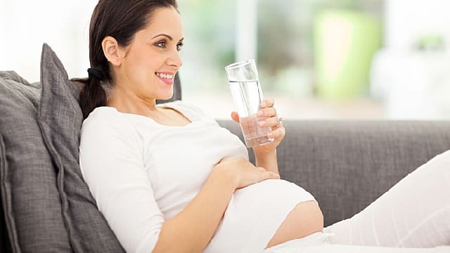 a pregnant woman sitting on the sofa smiling while holding a glass of water