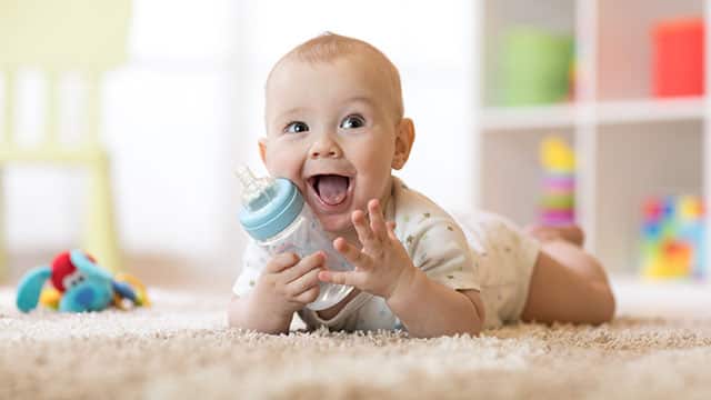 A baby boy is holding a bottle while laying on a bed