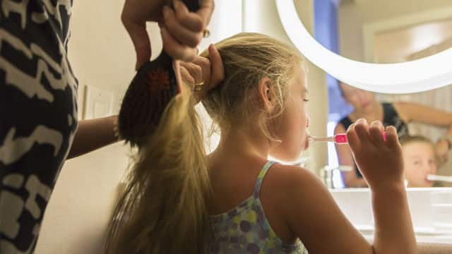 mother brushing her daugher hair while the girl is brushing her teeth with Colgate toothbrush