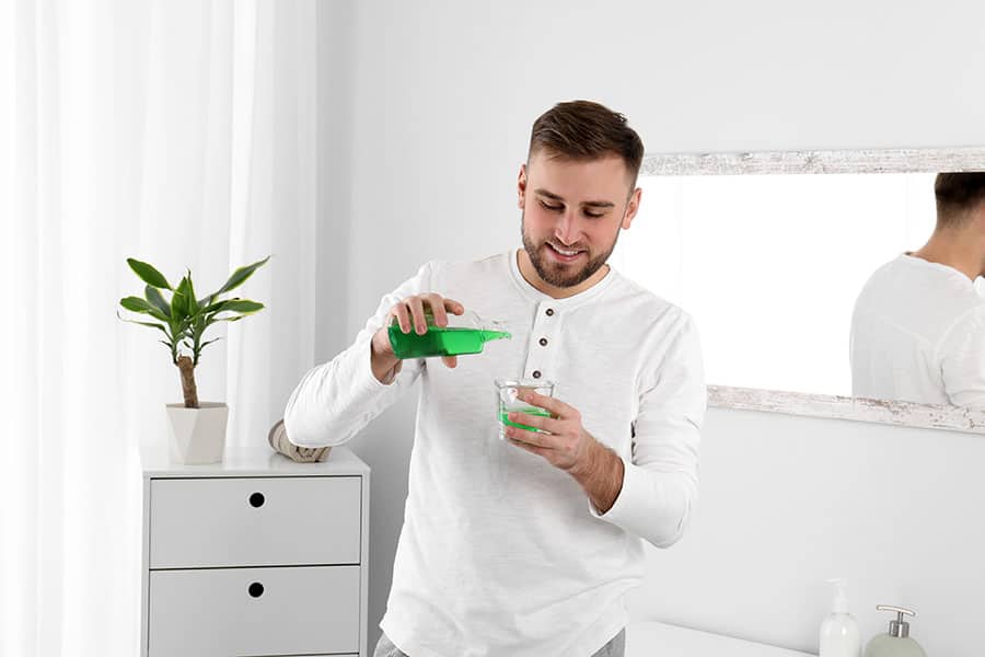 Young man with beard, wearing a white long sleeve shirt and pouring a green colored mouthwash into a small cup inside of a bathroom