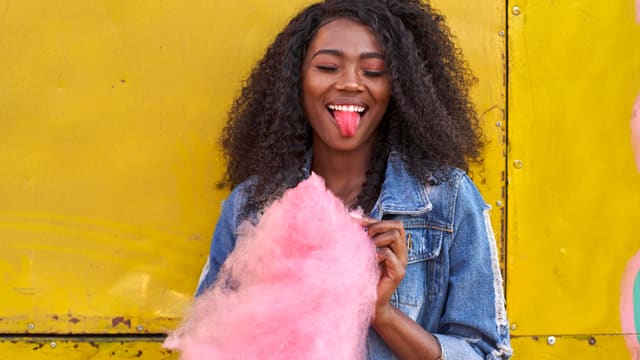 Portrait of smiling young woman in denim jacket with pink cotton candy sticking out tongue
