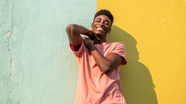 portrait of a young man looking at the camera as he leans on a light blue/yellow wall.