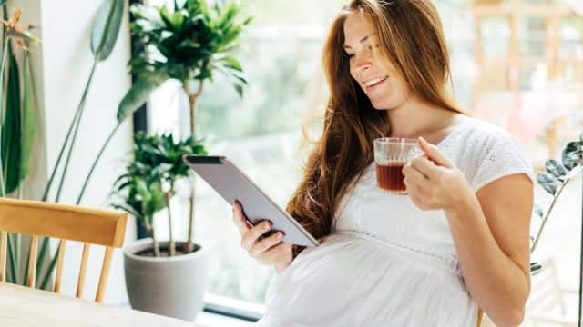 pregnant woman drinking from her cup while looking at her tablet
