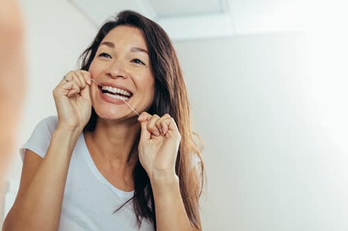 woman smiling while using a dental floss in front of her bathroom mirror