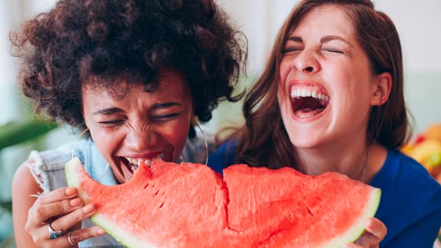 Two woman laughing while eating a large slice of watermelon