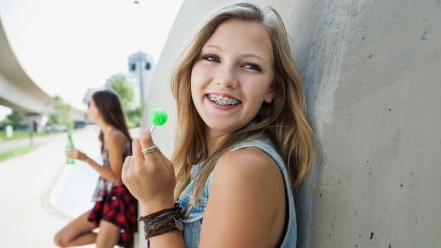 Smiling teenage girl with braces enjoying a lollipop with her friend in the background