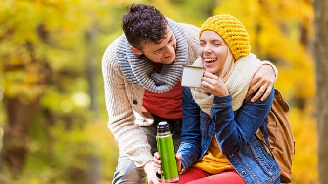 a man and a woman smiling while sitting down outdoor enjoying a cup of tea