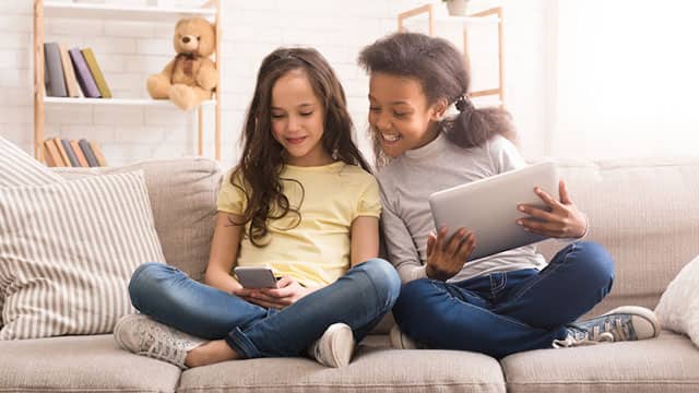 two young girls on a sofa playing on their tablet and phone