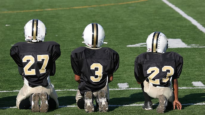 three boys kneeling wearing football gear in the football field watching the game
