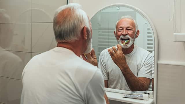 older man brusthing teeth in bathroom mirror