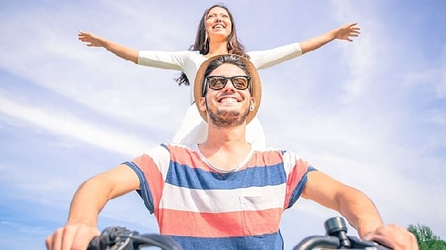 Man smiling riding a bicycle with woman smiling standing behind